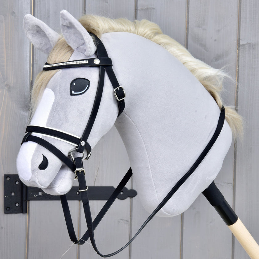 Hobby Horse di peluche con criniera bianca, briglia nera e una striscia bianca sul muso, fissato a un bastone di legno e posizionato davanti a uno sfondo in legno grigio.
