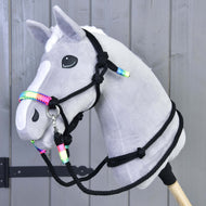 Hobby Horse grigio in peluche di Hobby Horse LarDen con lista bianca e criniera bianca, che indossa la cavezza Set Lunghina arcobaleno, montato su un bastone di legno davanti a pannelli in legno grigio.