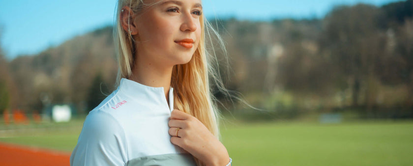 A young woman with long blonde hair stands outdoors on a sunny day, wearing a white athletic jacket and looking thoughtfully into the distance, perhaps reflecting on her latest Hobby Horsing adventure, with a blurred grassy field and trees in the background.