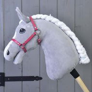 Hobby Horse LarDen in peluche grigio con criniera bianca intrecciata, occhio nero e Cavezza lampone (taglia M), fissato a un bastone di legno, davanti a una parete in legno grigio a pannelli.