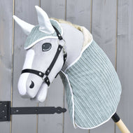 The Set Manchester Little Green by Hobby Horse LarDen features a gray and white hobby horse with a soft fabric head, black bridle, ear bonnet, and light blue-green neck cover on a wooden stick, shown against a wooden fence.