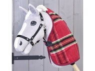 Un giocattolo Hobby Horse grigio di Hobby Horse LarDen, che indossa la Coperta da scuderia Christmas a quadri rossi e una briglia nera, montato su un bastone di legno davanti a uno sfondo di pannelli in legno chiaro.