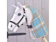 Un Hobby Horse in peluche di Hobby Horse LarDen, con muso grigio e bianco, briglia nera e Coperta da scuderia Scottish Cube in scozzese blu, su un bastone davanti a una parete di legno chiaro.