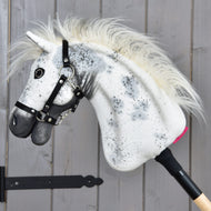 Hobby Horse Aspen con Cavezza nera, taglia S, di Hobby Horse LarDen, con testa in peluche bianca decorata da macchie nere, criniera soffice e cavezza nera, montato su un bastone di legno e mostrato su uno sfondo di parete in legno grigio.