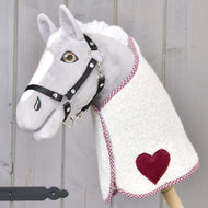 The Hobby Horse LarDen plush hobby horse has a white and gray face, black bridle, and comes with a Blanket cube red fleece blanket featuring a red heart patch, mounted on a wooden stick against light wood.
