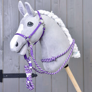 Set Start Viola (cavezza + redini a collare + lunghina + redini) di Hobby Horse LarDen su Hobby Horse di peluche con testa grigia, criniera bianca e lista bianca, montato su bastone in legno davanti a una parete in legno grigio.