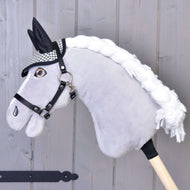 Hobby Horse LarDen con Cuffia inglese nera a quadri, Hobby Horse in peluche con muso grigio, criniera in filato bianco, finimenti neri e cuffia a quadri per protezione dagli insetti, montato su un bastone di legno contro una parete grigia.