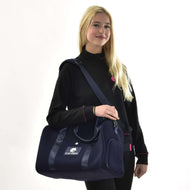 A smiling woman with long blonde hair, in a black jacket with pink trim, stands against a white background holding the Sports bag LarDen by Hobby Horse LarDen—a navy blue bag with adjustable strap, ideal for everyday use.