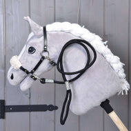 The Hobby Horse LarDen Set Basic features a plush hobby horse with a white mane, black bridle with reins, and black halter on a wooden stick, shown leaning against gray wooden panels.