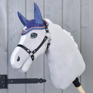 The Hobby Horse LarDen Ear Bonnet Crocheted Blue Sport is shown on a plush hobby horse with a white face and black bridle, mounted on a wooden stick in front of a gray panel door with a black hinge.