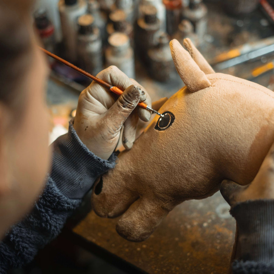 Una persona con i guanti usa un pennello per dipingere con cura l’occhio di un peluche Hobby Horse marrone, con vasetti di vernice visibili sullo sfondo sfocato.