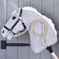 Hobby Horse - Corda guida lavorata a maglia Double Latte di Hobby Horse LarDen, cavallo su bastone grigio con briglia nera e redini intrecciate, esposto davanti a una parete di legno grigia.