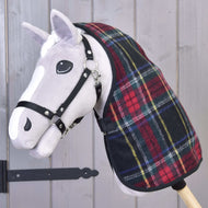 Un Hobby Horse in peluche con muso bianco e briglia nera indossa la Coperta da scuderia Edinburgh di Hobby Horse LarDen a quadri rossi e verdi, posato davanti a una parete di legno grigia con una cerniera metallica nera visibile.
