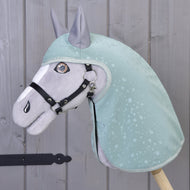 Hobby Horse LarDen Impermeabile Greeny su cavallo in peluche, vestito con un impermeabile verde con motivo a gocce d'acqua e briglia nera, accanto a una parete in legno grigio. I dettagli ricamati e le orecchie dritte offrono divertimento con ogni tempo.