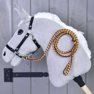 The Hobby Horse LarDen's "Hobby Horse - Knitted Lead Rope Pomello" features a plush gray hobby horse with a white mane, black bridle, and an orange-black knitted lead rope with carabiner, mounted on a wooden wall.