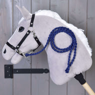 A gray plush Hobby Horse - Knitted Lead Rope Royal Blue by Hobby Horse LarDen, featuring a white mane, black bridle, and royal blue hand-knitted lead rope, is mounted on a wooden wall with its stick secured by a black metal bracket.