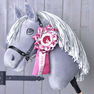 Nastro Hobby Horse LarDen Rosa di Hobby Horse LarDen su un Hobby Horse grigio con criniera in filato bianco, briglia nera e coccarda da dressage rosa e bianca sulla testa, mostrato su sfondo in legno chiaro.