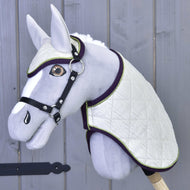 Set Leonardo di Hobby Horse LarDen con Hobby Horse di peluche grigio, coperta bianca trapuntata, cuffia antimosche coordinata, briglia nera e redini su un bastone di legno, mostrato davanti a una parete di legno grigia.