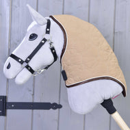 Primo piano della Copertura collo Bonanza di Hobby Horse LarDen: un Hobby Horse grigio con briglia nera e coperta trapuntata beige con chiusure sicure, montato su un bastone di legno davanti a una parete in legno grigio.
