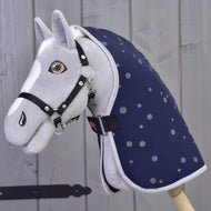 Hobby Horse in peluche grigio di Hobby Horse LarDen con briglia nera e Coperta da scuderia Night Sky, montato su un bastone di legno davanti a una parete in legno chiaro.