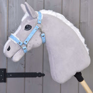 Un cavallo giocattolo di peluche grigio su bastone con criniera bianca è mostrato con la Hobby Horse LarDen Cavezza con pelliccia - similpelle Pastell Blue, appoggiato a una parete di legno a pannelli con cerniere metalliche nere.