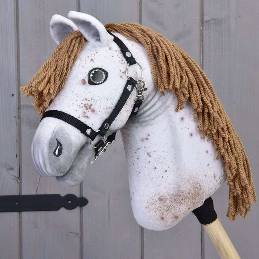 Hobby Horse Rufus con cavezza nera di Hobby Horse LarDen con testa artigianale in tessuto bianco maculato, criniera in filato marrone, occhi a bottone e cavezza nera su bastone di legno, su sfondo in legno grigio.