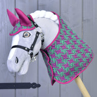 Un Hobby Horse LarDen grigio davanti a una parete di legno grigio chiaro indossa il Set Coco: set coperta con motivo pied-de-poule rosa e verde, cuffietta rosa coordinata e briglia nera.