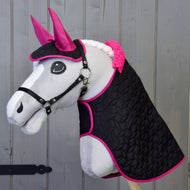 The Hobby Horse LarDen "Set Thermo Black with Fur" is a plush hobby horse with a white face, pink ears, black bridle, and a braided white mane, shown against a wooden background. It features a black quilted blanket trimmed in bright pink.