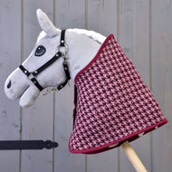 The Hobby Horse LarDen "Set festive Bordeaux" features a gray plush hobby horse with a black bridle and a burgundy-pink houndstooth blanket, mounted on a wooden stick for charming decor against a gray wooden backdrop.