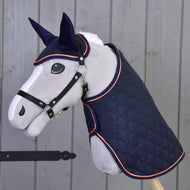 Hobby Horse LarDen "Set Winner" con testa di cavallo imbottita su bastone, vestita con coperta trapuntata blu navy e cuffietta per orecchie coordinata con bordo arancione, posata davanti a una parete in legno.