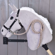 The Hobby Horse LarDen Hobby Horse - Knitted Lead Rope Almond features a light gray plush horse with a white mane, black bridle, hand-knitted almond lead rope, and is displayed on a wooden stick against a gray wooden wall.