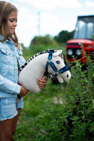 Ragazza con giacca di jeans accanto a una siepe con Hobby Horse Aurora con Cavezza nera di Hobby Horse LarDen, testa in peluche bianco crema e criniera in filato a righe nere e bianche, con Cavezza LarDen Blue foderata di pelliccia e trattore rosso sfocato sullo sfondo
