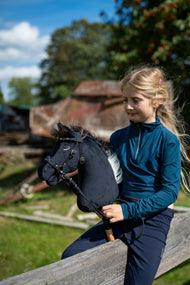 Ragazza bionda con maglia verde petrolio seduta su una staccionata di legno in una fattoria soleggiata con Hobby Horse Bond con Cavezza nera di Hobby Horse LarDen, testa in peluche nero e criniera in filato marrone scuro, con Briglia senza morso decorata