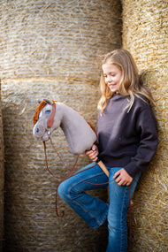 Ragazza sorridente con felpa marrone scuro appoggiata a balle di paglia rotonde con Hobby Horse Chanell con Cavezza nera di Hobby Horse LarDen, testa in peluche grigio, occhio blu e criniera in filato rosso rame, con Briglia Western a un orecchio completa