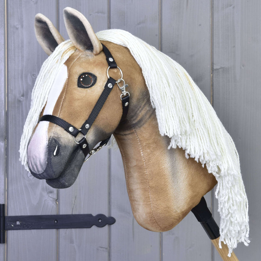Hobby Horse Connie con Cavezza Nera di Hobby Horse LarDen, con realistico design marrone chiaro e bianco, lunga criniera bianca in filato e montato su un bastone davanti a una parete di legno grigio.