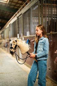 Ragazza con giacca di jeans che conduce Hobby Horse Dancing Lady con Cavezza nera di Hobby Horse LarDen nel corridoio di una scuderia, testa in peluche crema, criniera e ciuffo in filato chiaro, con Cavezza nera in similpelle con pelliccia nera completa e Redini nere