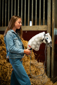 Ragazza con giacca di jeans davanti a balle di paglia in una scuderia con Hobby Horse Elektra con Cavezza nera di Hobby Horse LarDen, testa in peluche bianco pomellato, occhio blu e criniera in filato bianco, con Cavezza Graphite Full con pelliccia in similpelle e Lunghina lavorata a maglia