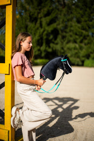 Ragazza con maglia rosa appoggiata a un'ala di ostacolo gialla con Hobby Horse Freya con Cavezza nera di Hobby Horse LarDen, testa in peluche nero, lista bianca e criniera in filato nero, con Briglia turchese completa senza Nasello