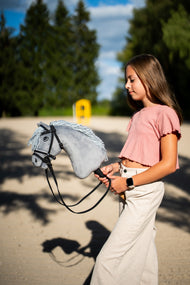 Ragazza con maglia rosa che conduce Hobby Horse Grace con Cavezza nera di Hobby Horse LarDen in un campo in sabbia, testa in peluche grigio e criniera in filato grigio chiaro, con Briglia De Luxe completa con due frontalini