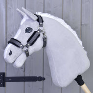 Una Cavezza con pelliccia - similpelle Graphite Full di Hobby Horse LarDen è montata su un bastone di legno e appoggiata a una parete di legno grigio chiaro, con testa di peluche grigia, criniera bianca, briglia nera e cavezza in similpelle.