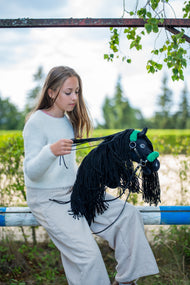 Una ragazza con maglione chiaro siede sulla staccionata del paddock raccogliendo le redini dell'Hobby Horse Maximus con Cavezza nera di Hobby Horse LarDen, testa in peluche nero e lunga criniera in filato nero, qui mostrato con cavezza nera in similpelle con pelliccia verde e redini nere in similpelle