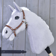 Hobby Horse LarDen grigio di peluche con criniera bianca e marcature sul muso, con Cavezza - similpelle marrone, montato su un bastone di legno ed esposto davanti a una parete di legno grigia.
