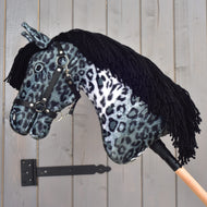 Hobby Horse Picasso con cavezza nera di Hobby Horse LarDen, con testa in peluche dal mantello maculato grigio e nero ispirato all'Appaloosa, cavezza nera e criniera in filato nero, montato su un bastone di legno e mostrato davanti a una parete di legno grigia.