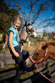 Una ragazza bionda con top turchese è seduta su una recinzione di legno del paddock con il Hobby Horse Piccolino con Black Halter di Hobby Horse LarDen, testa in peluche beige chiaro e criniera in filato scuro, con un pony sauro dietro di lei e la briglia nera Complete in similpelle