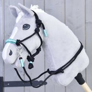 Il Set corda Turquoise Hobby Horse LarDen è mostrato su un Hobby Horse grigio di peluche con criniera bianca, briglia nera e dettagli turchesi, montato su un bastone davanti a una parete di legno grigio.