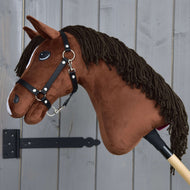 Hobby Horse Rocky con Cavezza Nera di Hobby Horse LarDen, cavallo a bastone in peluche marrone, mostrato accanto a una parete in legno grigio.