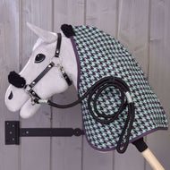 Set Harmony Green di Hobby Horse LarDen: un Hobby Horse di peluche con cavezza nera, coperta da scuderia verde e viola a motivo pied-de-poule e corda nera arrotolata, montato su un supporto da parete in legno davanti a una parete di legno grigia.