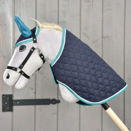 The Hobby Horse LarDen Set Supreme Turquoise features a gray plush stick horse with blue ears, a black bridle, and a quilted navy blanket with teal trim, mounted on a wooden pole and set against a light wood wall.