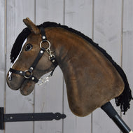 Hobby Horse Sundance con cavezza nera di Hobby Horse LarDen, con design in peluche, criniera in filato nero, briglia e redini, che richiama l'aspetto vivace di uno stallone Purosangue inglese, montato su un bastone e mostrato davanti a una parete di assi di legno chiaro.