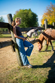 Una ragazza con top marrone e jeans si appoggia a un palo di recinzione di legno con il Hobby Horse Thor con Black Halter di Hobby Horse LarDen, testa in peluche grigio e criniera in filato nero, con un pony al pascolo nel paddock e la briglia nera Complete con diamanti